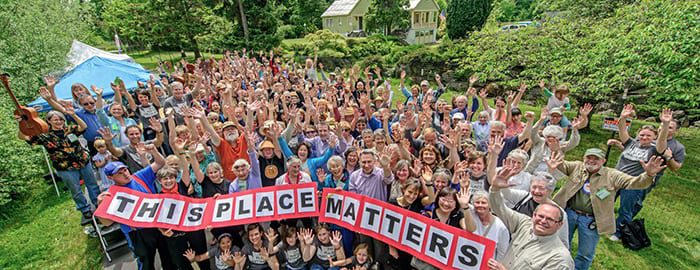 A large group of people with their hands in the air. A line of people at the front of the gathering carries a red, white, and black sign that reads "THIS PLACE MATTERS"