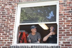 Two men seen through an white, open window in a brick building. The man on the left observes the man on the right as he makes adjustments to the window sill.