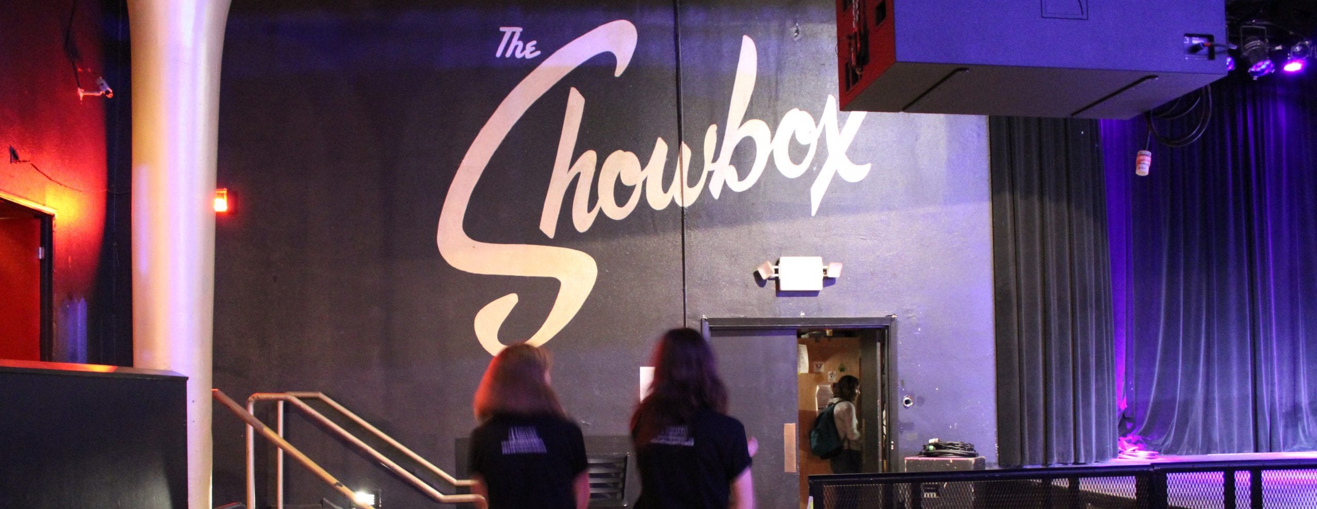 Two women in "Save The Showbox" shirts stand beside each other, looking a black wall inside The Showbox. On the wall are the words "The Showbox" in large, white letters.