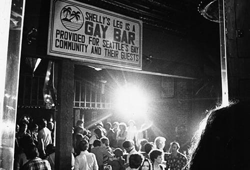 Bar patrons socialize underneath a sign that reads "Shelly's Leg is a gay bar provided for Seattle's gay community and their guests."