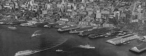 An old photo of Seattle's waterfront, with several ships sailing in and out of various piers..