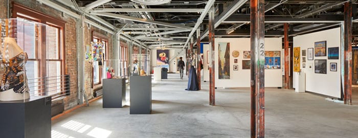 A gallery space with an exposed brick wall to the left, flanked by objects in glass containers on pedestals. To the right, walls covered in artwork including paintings and photographs.