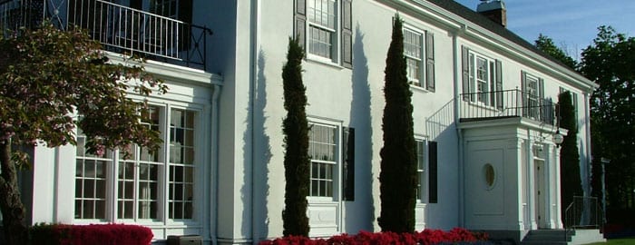 A close-up of the Admiral's House, a white house lined with red flowers along the exterior. Four tall trees stand in front of the house.