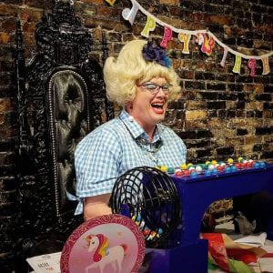 Sylvia O'Stayformore, a drag queen in a blonde wig and blue and white checkered dress, calls out bingo numbers. In front of her are a collection of colorful bingo balls.