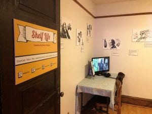 An open door labeled "Shelf Life Community Story Project" leads to a room adorned with sketches of storytellers. A computer in the corner has headphones for listening to the stories.